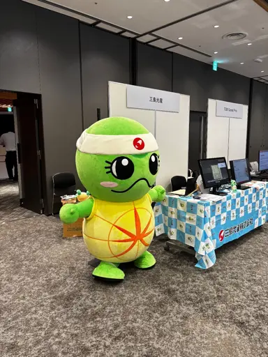 Big inflatable turtle with a plush texture standing next to a booth at a convention. The turtle is sanding on their back legs , is wearing a white headband and shows a dead-serious expression! 