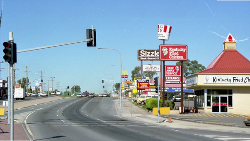 A photo of a stroad with 4 lanes in the oncoming direction. On the side is visible a number of stores and restaurants and advertising signs, including for KFC, Sizzler, McDonalds, and Sony Betamax.